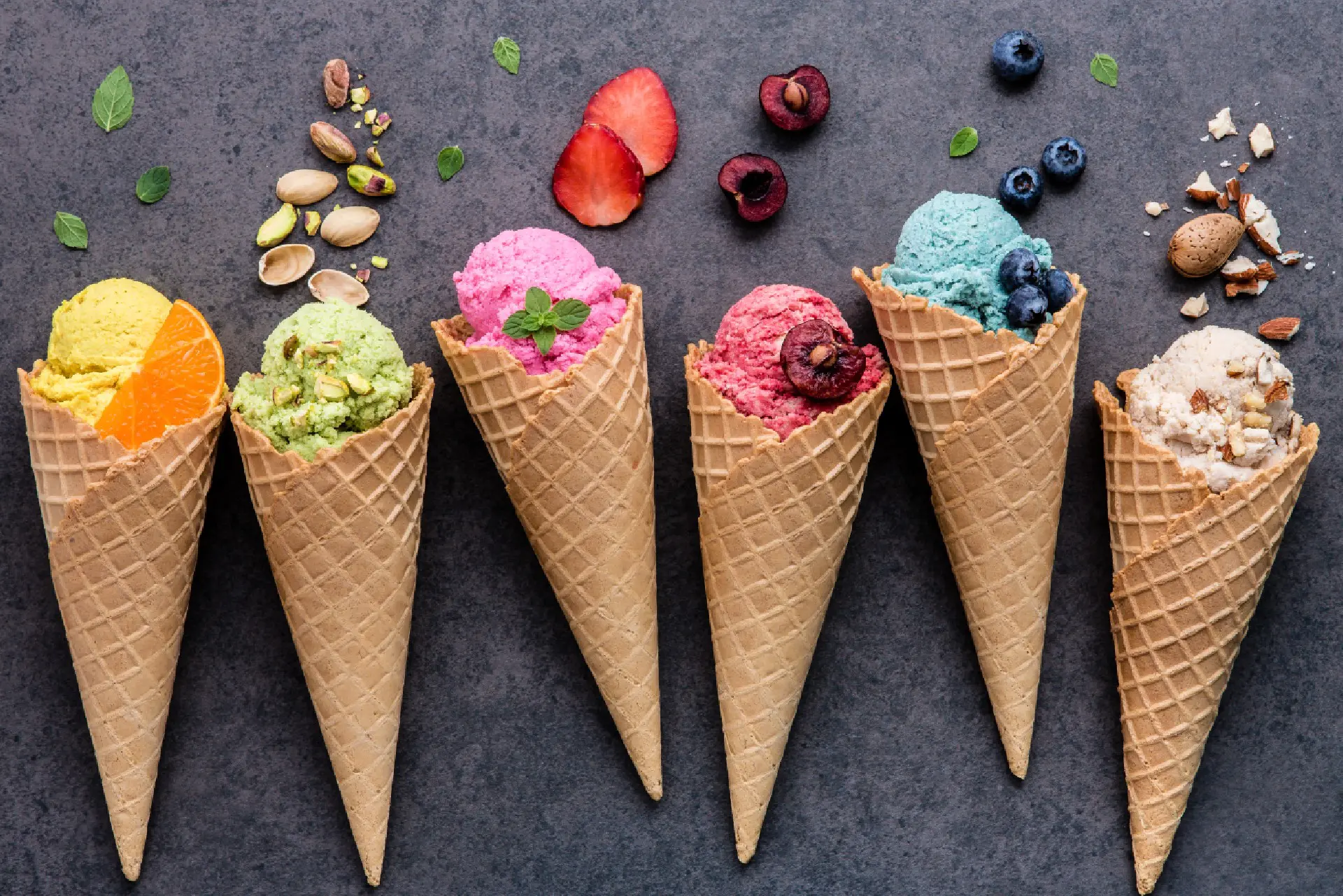 Six waffle cones with various colorful ice cream scoops and different toppings are arranged on a textured gray surface.