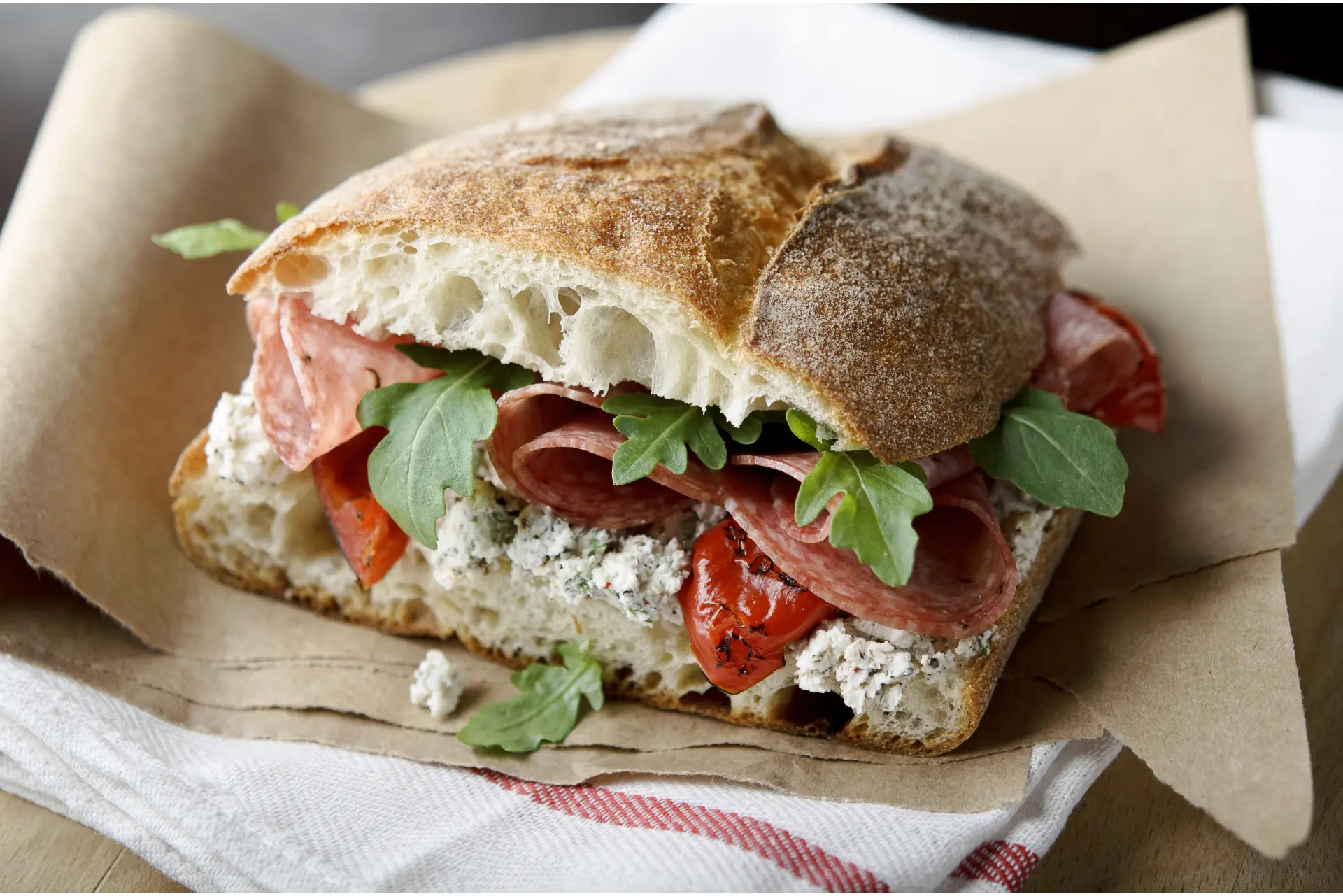 A sandwich with rustic bread, salami, arugula, peppers, and herb cream cheese is placed on brown paper over a white cloth with a red stripe.