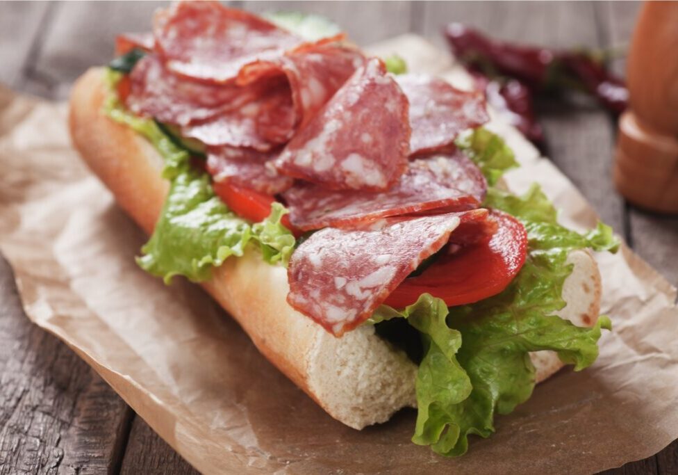 A sandwich on brown paper containing slices of salami, lettuce, and tomato on a wooden table.