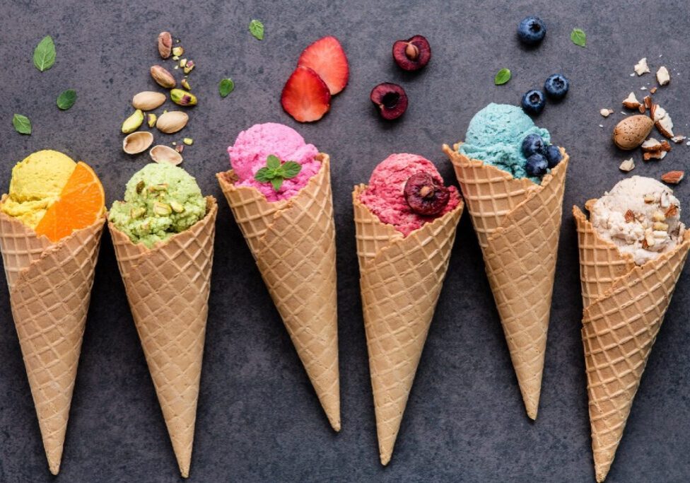 Six waffle cones with various colorful ice cream scoops and different toppings are arranged on a textured gray surface.