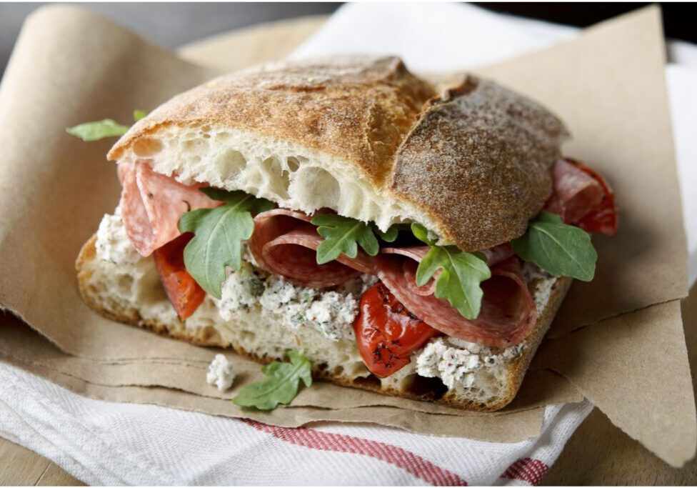 A sandwich with rustic bread, salami, arugula, peppers, and herb cream cheese is placed on brown paper over a white cloth with a red stripe.